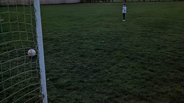 A child stands in a football field near a goalpost at sunset, with trees silhouetted in the background.