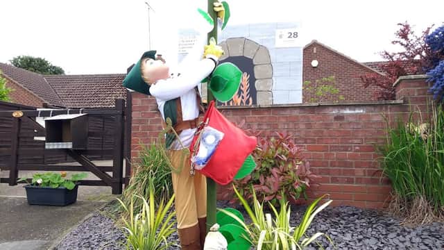 A scarecrow figure climbing a post, wearing a green hat, with a red bag and surrounded by plants in Wetwang.