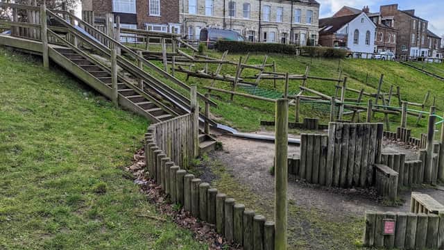 Wooden play structures and a grassy hill, with houses in the background under a partly cloudy sky.