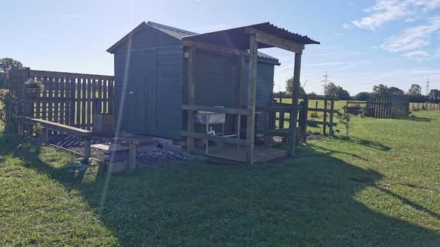 A wooden shed with a porch in a grassy field under a clear blue sky, surrounded by a wooden fence.