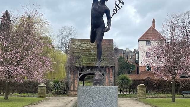 A bronze statue of a figure with a staff stands on a pedestal, surrounded by blooming trees and a pond.