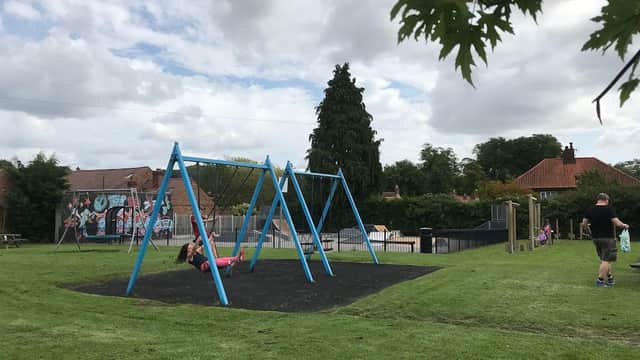 Colorful swings in a grassy park, with a child swinging and a man nearby, surrounded by trees and buildings.