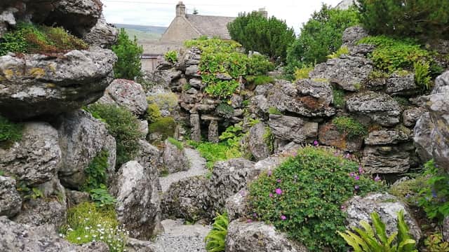 Lush greenery and stone formations create a serene pathway in the Aysgarth Edwardian Rock Garden, surrounded by buildings.