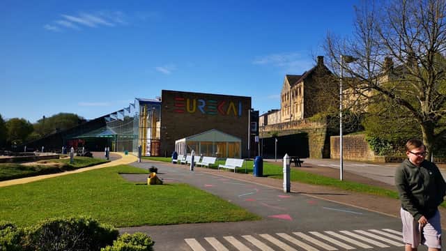 Pay & Display car park with colorful building, benches, and green space under a clear blue sky in Exley, Yorkshire.