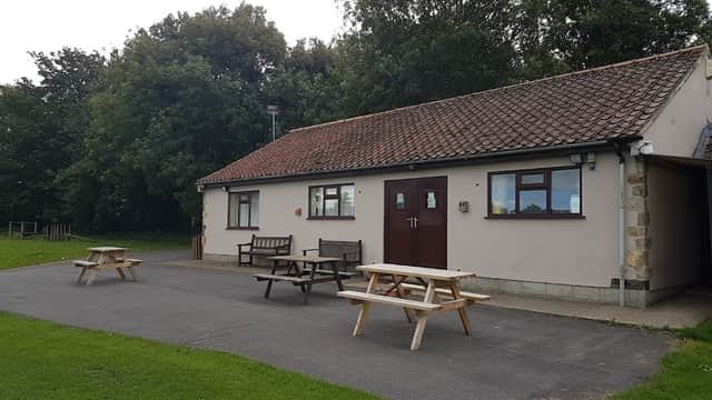 A sports club building with a brown door, picnic tables, and green grass, surrounded by trees in Slingsby, Yorkshire.