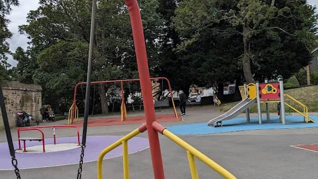 Colorful playground with swings, slides, and climbing structures, surrounded by trees and a cloudy sky.