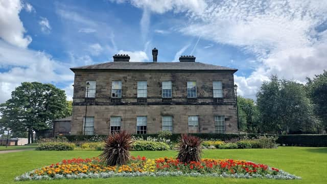 Historic stone building with large windows, surrounded by vibrant flower beds and lush green lawns under a blue sky.