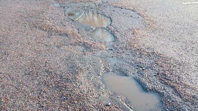 Potholes and puddles on a gravel parking lot with parked cars in the background, surrounded by trees.