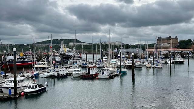 Harbor filled with boats and yachts under a cloudy sky, with buildings and hills in the background.
