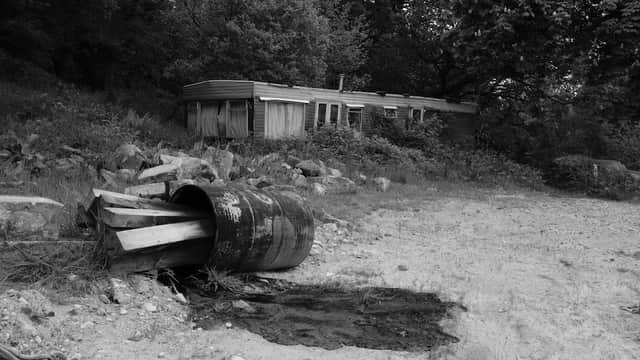 A derelict caravan surrounded by trees, with a rusty barrel and wooden planks in the foreground on a gravel path.