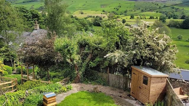 Lush green garden with beehives, wooden shed, and rolling hills under a partly cloudy sky in Thornton in Craven.