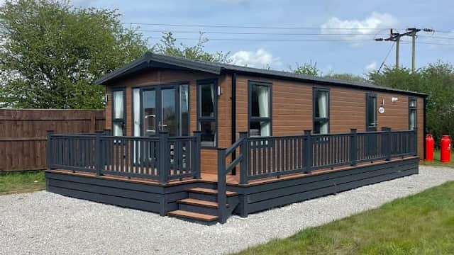 Modern holiday lodge with wooden exterior, deck, and gravel area, surrounded by greenery at The Meadows Holiday Park.