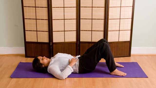 A woman lies on a yoga mat in front of a wooden screen, practicing the Alexander Technique in a calm setting.