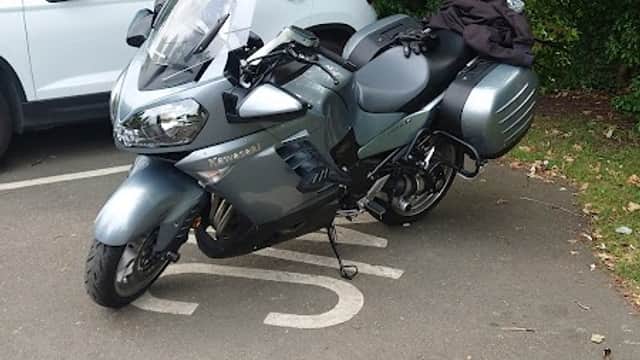 A gray motorcycle parked in a charging space, surrounded by greenery and a white vehicle in the background.