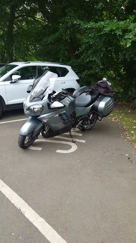 A gray motorcycle parked in a charging space, surrounded by greenery and a white vehicle in the background.