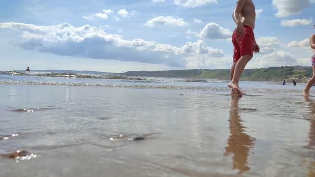 A child in red swim trunks walks along a sandy beach with gentle waves and a cloudy sky in the background.