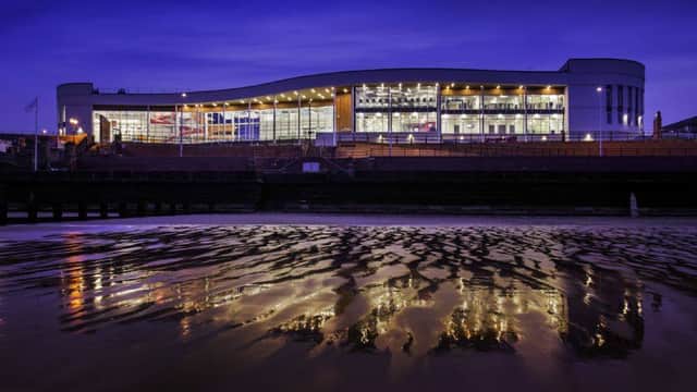 Modern spa building with large windows reflecting on wet sand at dusk, illuminated by warm lights.