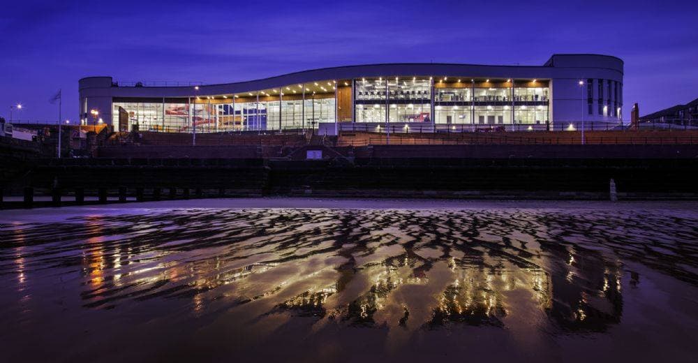 Modern spa building with large windows reflecting on wet sand at dusk, illuminated by warm lights.