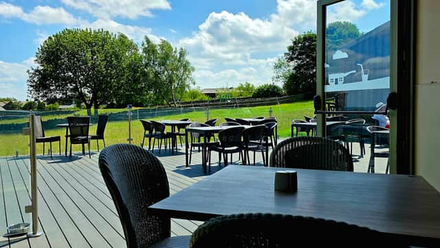 Outdoor seating area with black chairs and tables, overlooking green fields under a blue sky with fluffy clouds.
