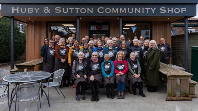 A large group of smiling volunteers in aprons stands outside the Huby & Sutton Community Shop, with tables and chairs nearby.