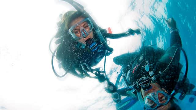 Two divers underwater, wearing scuba gear and masks, surrounded by bubbles and clear blue water.