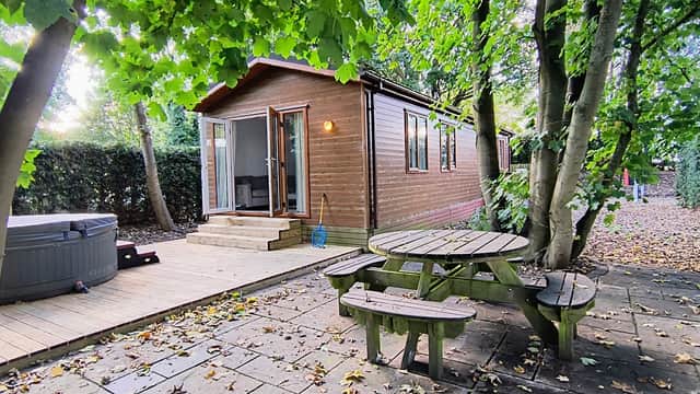 Wooden cabin surrounded by trees, with a hot tub and picnic table on a stone patio.