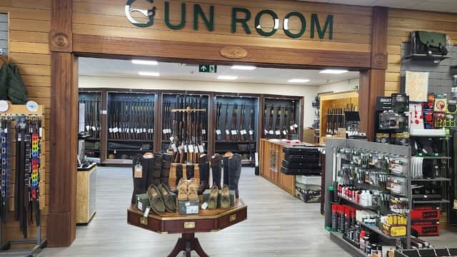 Gun Room display with various firearms, accessories, and footwear in a wooden-paneled shop interior.