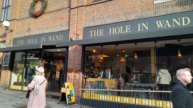 Shopfront with "The Hole in Wand" signage, festive wreath, people walking by, and a cozy interior visible through the windows.