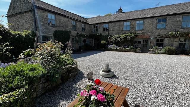 Charming stone cottages with a gravel courtyard, surrounded by greenery and flowers under a clear blue sky.