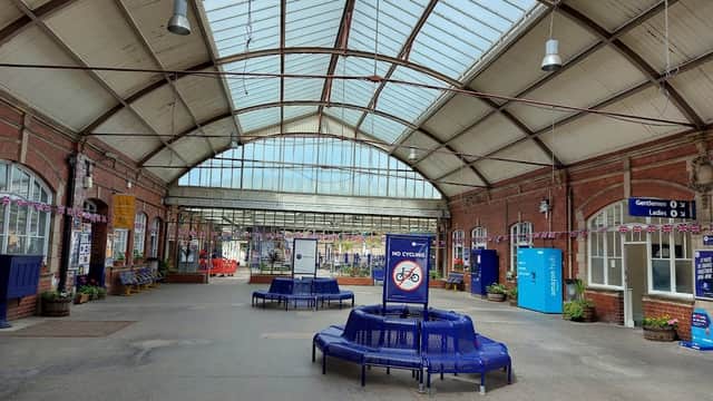Spacious railway station interior with high glass roof, blue seating, and signage, featuring colorful flower planters.