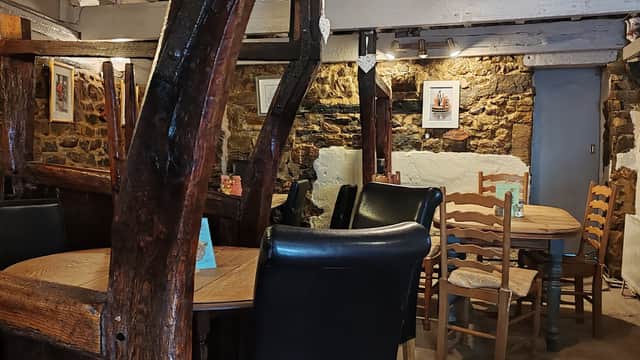 Cozy cafe interior with wooden beams, tables, chairs, and framed artwork on stone walls.