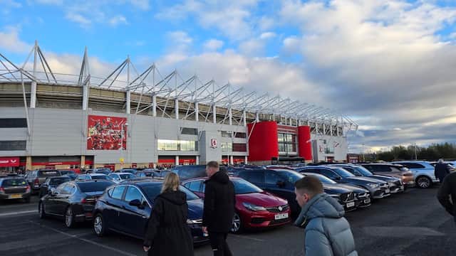 Busy car park with numerous parked cars and people walking, alongside a large stadium under a cloudy sky.
