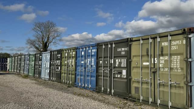 Colorful shipping containers lined up in a gravel yard under a partly cloudy sky, with a tree in the background.