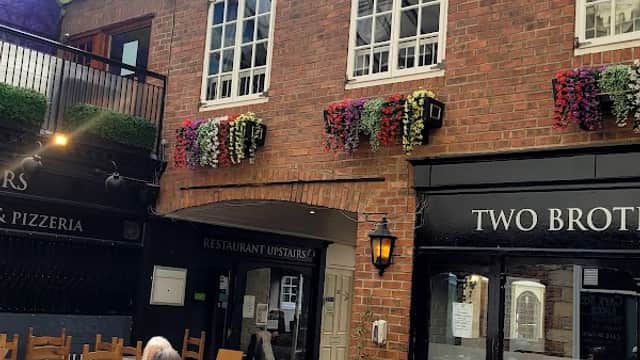 Brick walls adorned with flower baskets, glass ceiling, wooden chairs, and restaurant entrances in a cozy courtyard setting.