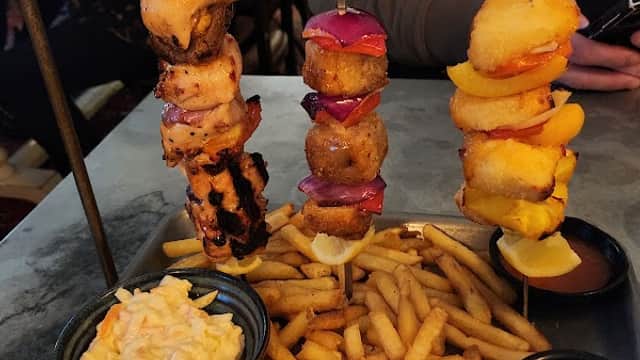 Skewers of grilled meats and vegetables served with coleslaw and fries on a metal tray, set in a busy restaurant.