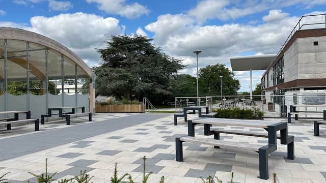 Modern corporate office building with a landscaped plaza, benches, greenery, and a large tree under a cloudy sky.