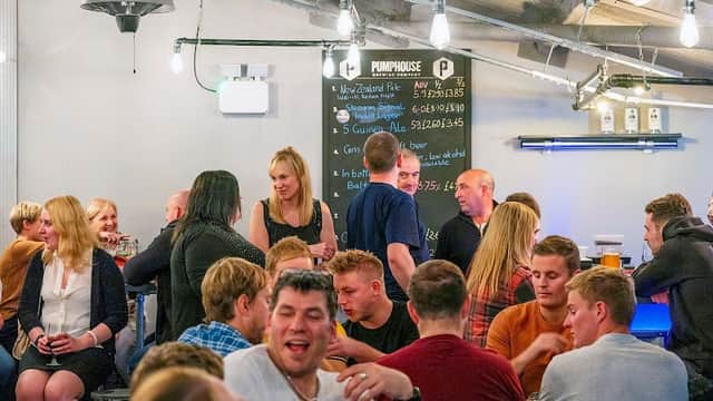 Lively brewery interior with patrons socializing, a bar, and a chalkboard menu in a modern, industrial setting.