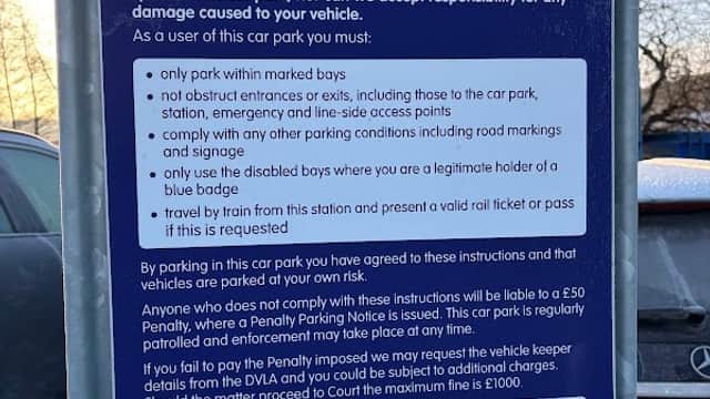 Sign detailing parking rules and regulations at Mirfield Station Car Park, with a fence and vehicles visible in the background.