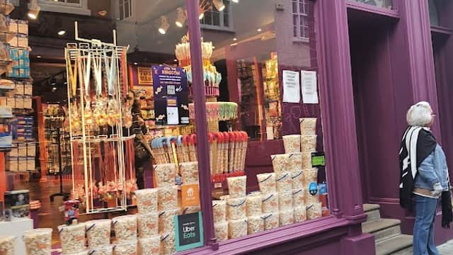Colorful shop window displaying various candies and snacks, with a woman standing nearby on a cobbled street.