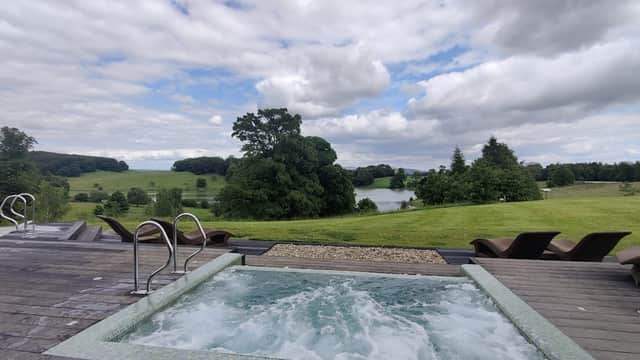 Hot tub on a wooden deck overlooking lush green fields and a tranquil lake under a cloudy sky.