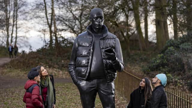 Visitors admire a towering sculpture of a man in a jacket, set in a wooded park landscape with a cloudy sky.