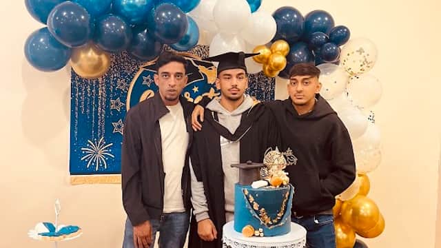 Graduation celebration with three young men posing by a decorated cake and dessert table, featuring blue and gold balloons.