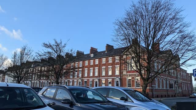Row of red-brick buildings under a clear blue sky, with parked cars in the foreground and bare trees nearby.