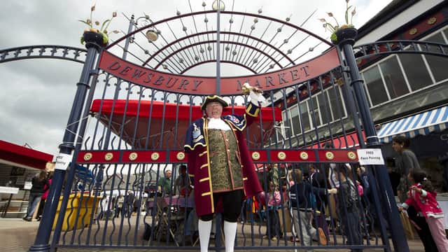 Town crier in traditional attire stands before ornate metal gate of Dewsbury Market, with bustling crowd in background.