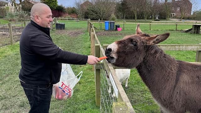A man feeding a donkey with a carrot at Clapham Holme Farm, surrounded by green fields and cloudy skies.