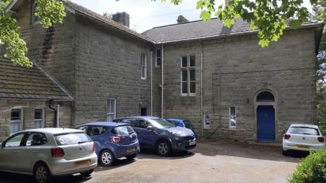Stone building with large windows, blue door, and a gravel parking area filled with several cars, surrounded by trees.