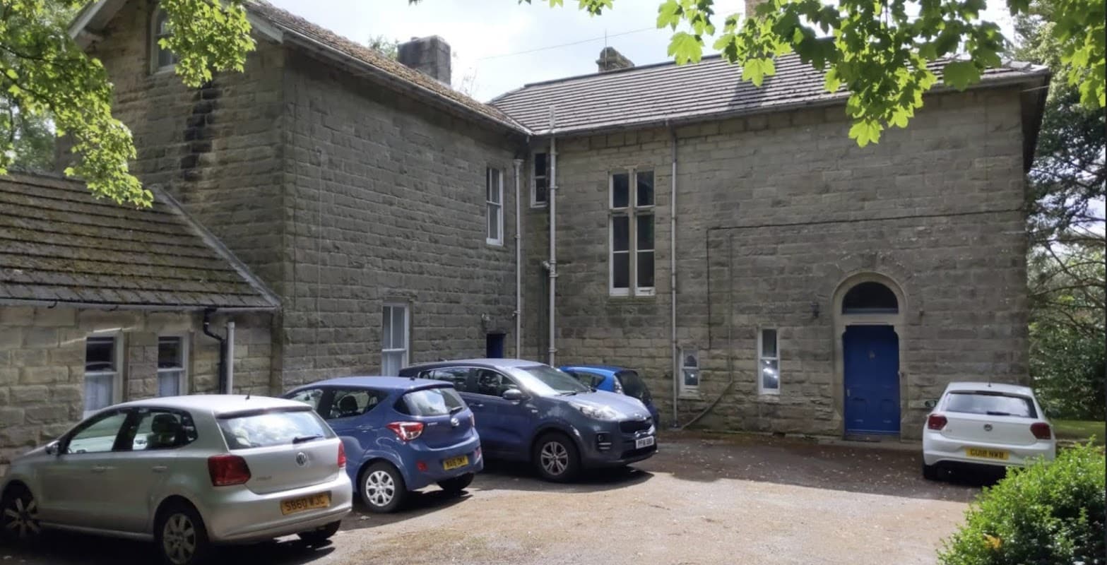 Stone building with large windows, blue door, and a gravel parking area filled with several cars, surrounded by trees.