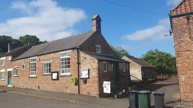 Brick building with large windows, green door, and recycling bins outside, set against a clear blue sky.