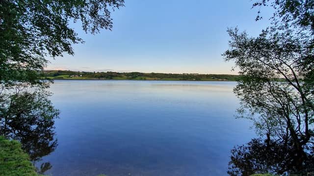 Serene view of Mixenden Reservoir surrounded by trees and a clear blue sky, reflecting calm waters.