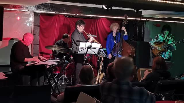 A jazz band performs in a dimly lit basement venue, with musicians on piano, saxophone, bass, and guitar.